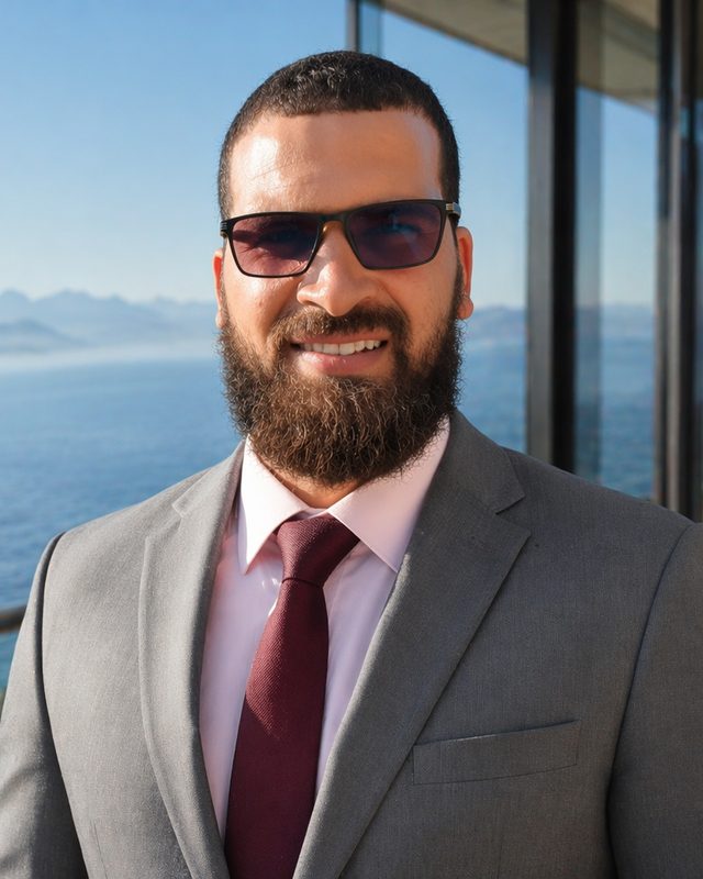 Portrait of Dr Rachid Khoumikham in a grey suit and burgundy tie, with the sea behind him