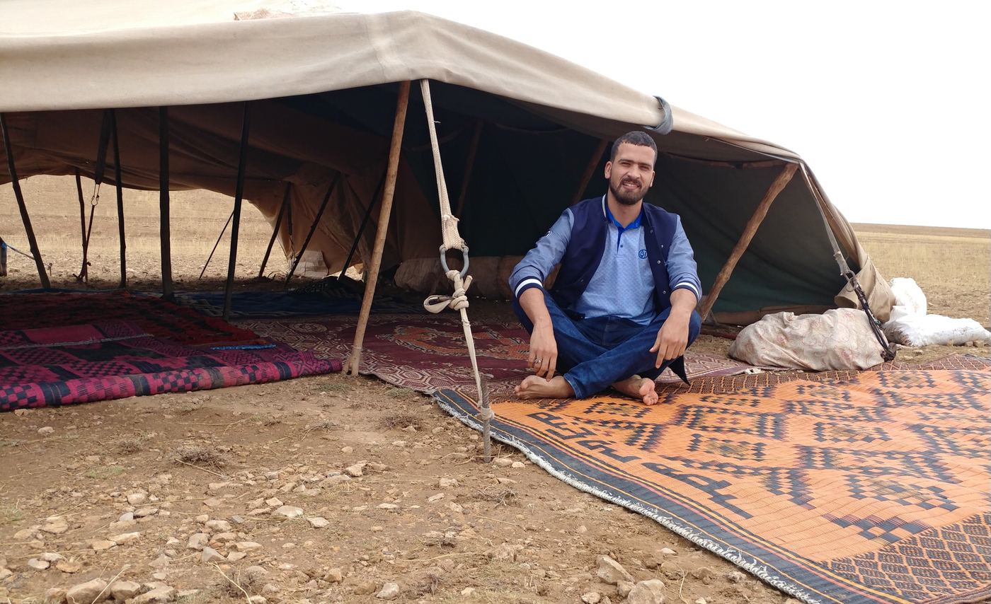 Rachid sitting cross-legged on a woven rug at the entrance of a nomadic tent on open steppe